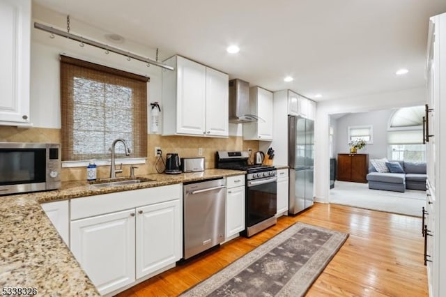 kitchen with light stone counters, a sink, white cabinets, appliances with stainless steel finishes, and wall chimney exhaust hood