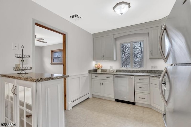 kitchen with a wainscoted wall, stainless steel appliances, a baseboard radiator, visible vents, and dark stone counters