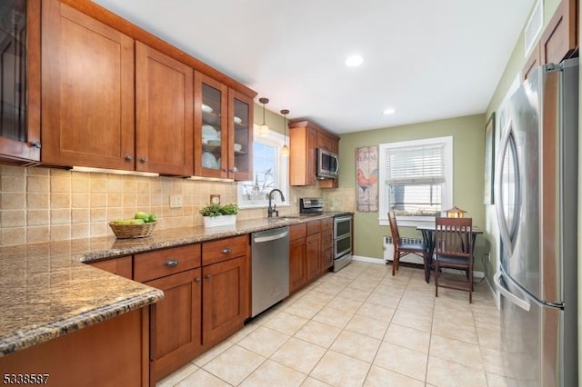 kitchen featuring light stone counters, stainless steel appliances, hanging light fixtures, brown cabinetry, and glass insert cabinets