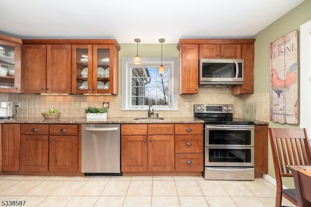 kitchen with light stone counters, decorative light fixtures, stainless steel appliances, glass insert cabinets, and a sink