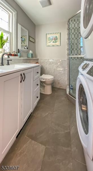 bathroom with a shower with door, tile walls, vanity, stacked washer / drying machine, and toilet