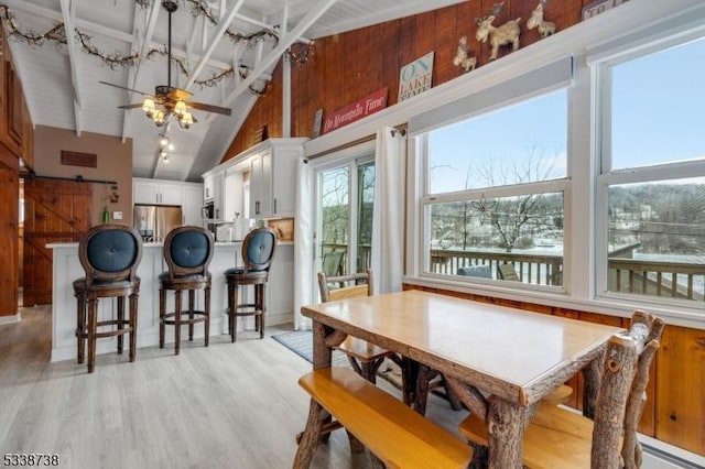 dining room featuring high vaulted ceiling, wooden walls, a ceiling fan, and light wood-style floors