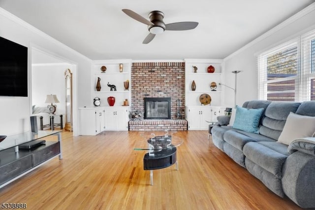 living area with light wood finished floors, built in features, a ceiling fan, crown molding, and a brick fireplace