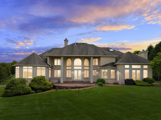 back of house at dusk with a patio, a lawn, a chimney, and stucco siding