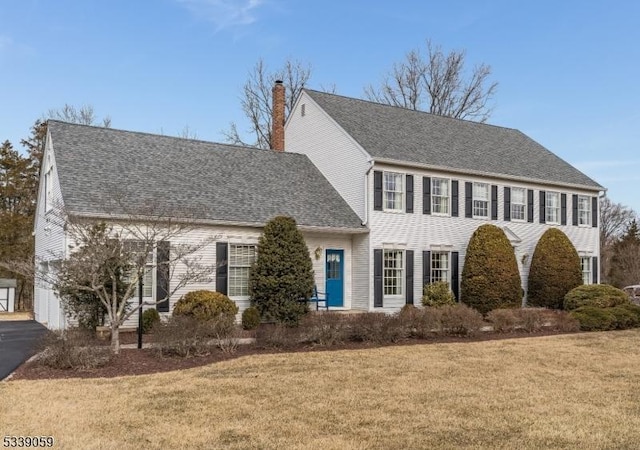 colonial home with roof with shingles, a front lawn, a chimney, and aphalt driveway