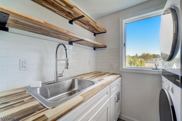 kitchen with butcher block countertops, sink, tasteful backsplash, stacked washer and clothes dryer, and white cabinets