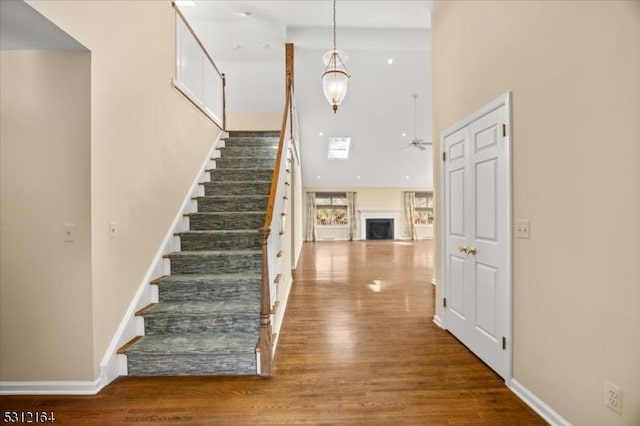 stairway featuring a high ceiling, a fireplace, wood finished floors, a ceiling fan, and baseboards