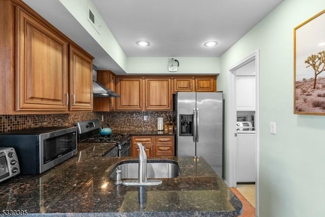 kitchen featuring stainless steel appliances, a sink, backsplash, brown cabinetry, and dark stone countertops