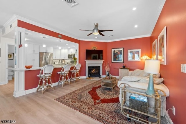 living room with light wood-type flooring, visible vents, a ceiling fan, and ornamental molding