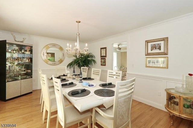 dining room featuring crown molding, wainscoting, a decorative wall, light wood-type flooring, and ceiling fan with notable chandelier