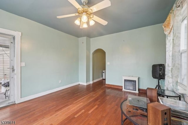 living room with arched walkways, a fireplace, wood finished floors, a ceiling fan, and baseboards