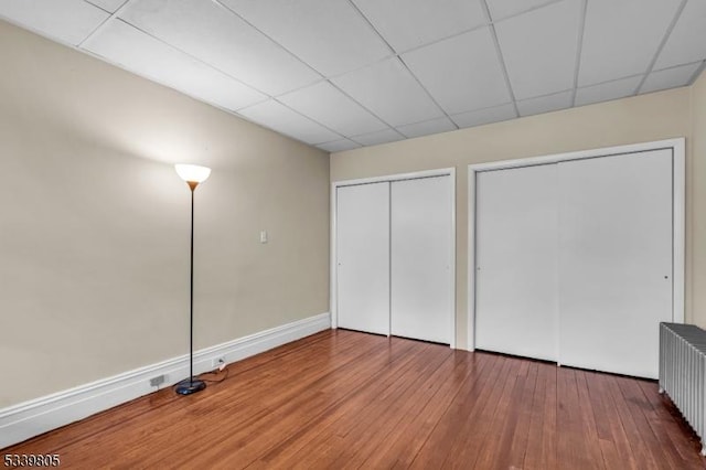 unfurnished bedroom with multiple closets, a paneled ceiling, radiator heating unit, wood finished floors, and baseboards