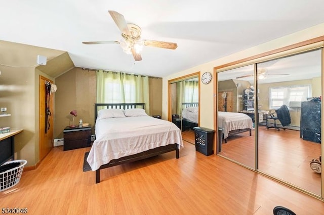 bedroom with a ceiling fan, lofted ceiling, light wood-type flooring, a baseboard heating unit, and multiple closets