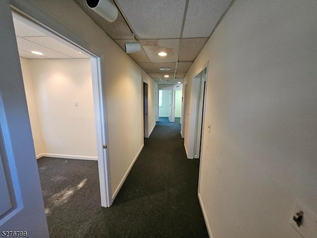 corridor featuring dark colored carpet, a drop ceiling, and baseboards