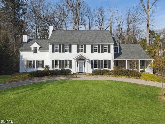 colonial house featuring roof with shingles, a chimney, and a front lawn