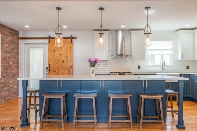 kitchen featuring light wood finished floors, a barn door, wall chimney exhaust hood, brick wall, and light countertops