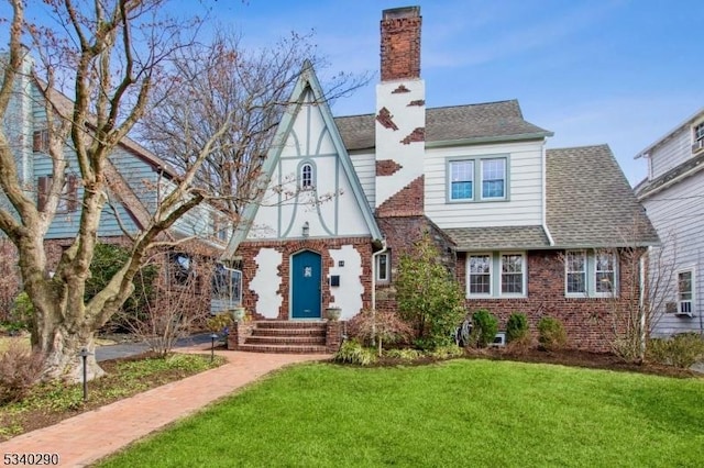 english style home with a shingled roof, a chimney, a front lawn, and brick siding