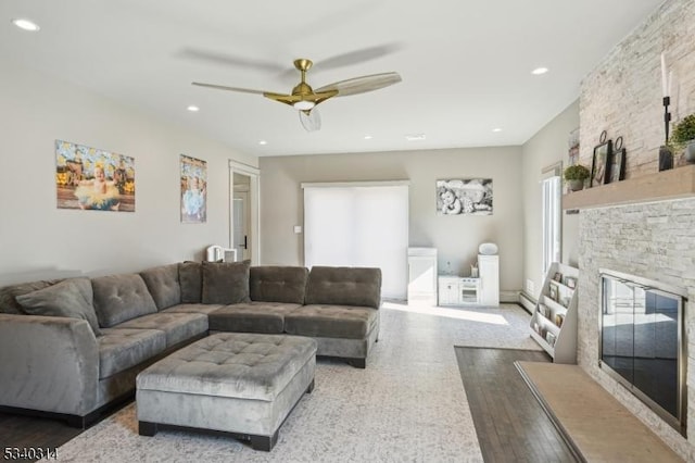 living area with a ceiling fan, wood finished floors, baseboard heating, a stone fireplace, and recessed lighting