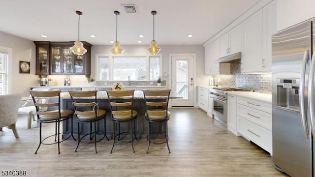 kitchen featuring backsplash, appliances with stainless steel finishes, glass insert cabinets, under cabinet range hood, and a kitchen bar