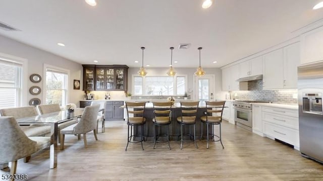 kitchen with under cabinet range hood, visible vents, appliances with stainless steel finishes, decorative backsplash, and glass insert cabinets