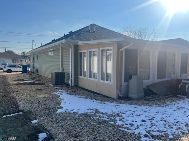 view of snowy exterior with central AC, roof with shingles, and a sunroom