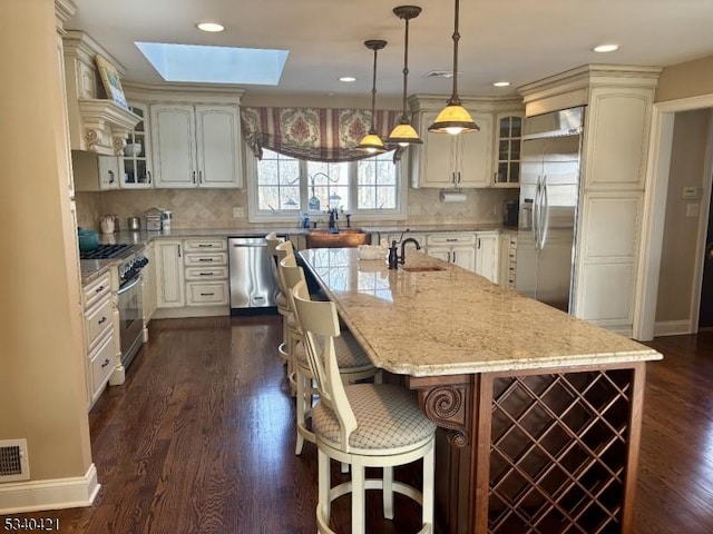 kitchen featuring a skylight, appliances with stainless steel finishes, glass insert cabinets, light stone counters, and a kitchen island with sink