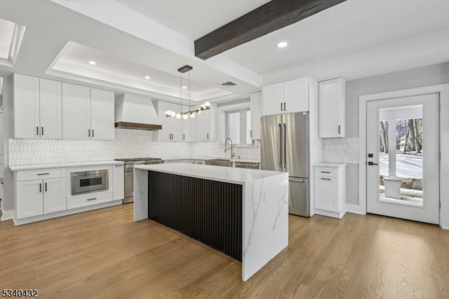 kitchen with a tray ceiling, light countertops, custom range hood, appliances with stainless steel finishes, and light wood-type flooring