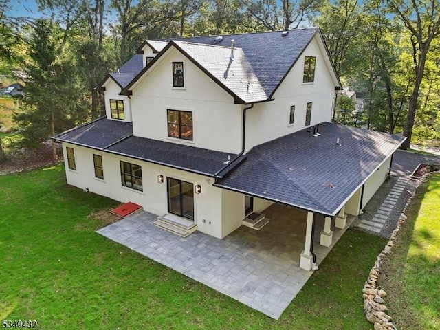 back of house with a shingled roof, stucco siding, a lawn, and a patio