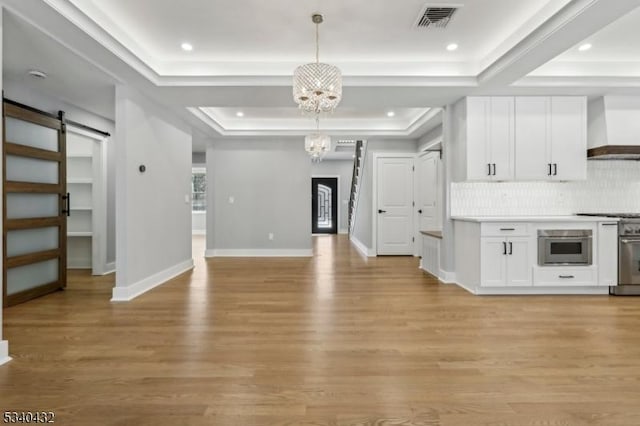 kitchen with a raised ceiling, a barn door, appliances with stainless steel finishes, a chandelier, and wall chimney exhaust hood