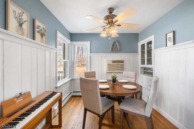 dining room featuring a wainscoted wall, a baseboard radiator, a wall mounted AC, light wood-style floors, and a ceiling fan