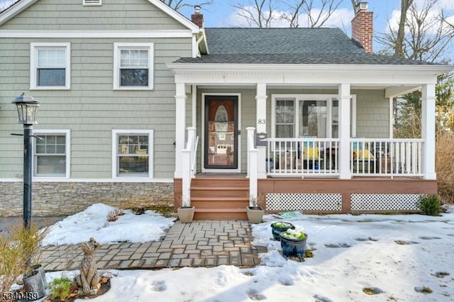 view of front of house with covered porch, a chimney, stone siding, and roof with shingles