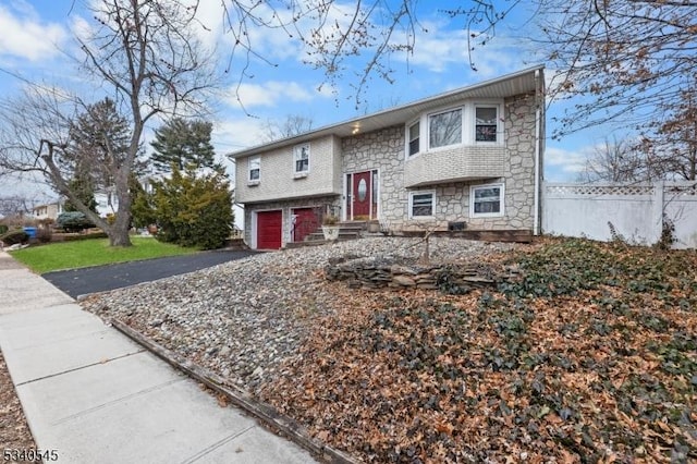 bi-level home featuring driveway, stone siding, an attached garage, and fence