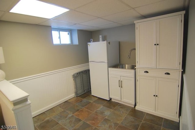 kitchen with white cabinets, a drop ceiling, a wainscoted wall, freestanding refrigerator, and a sink