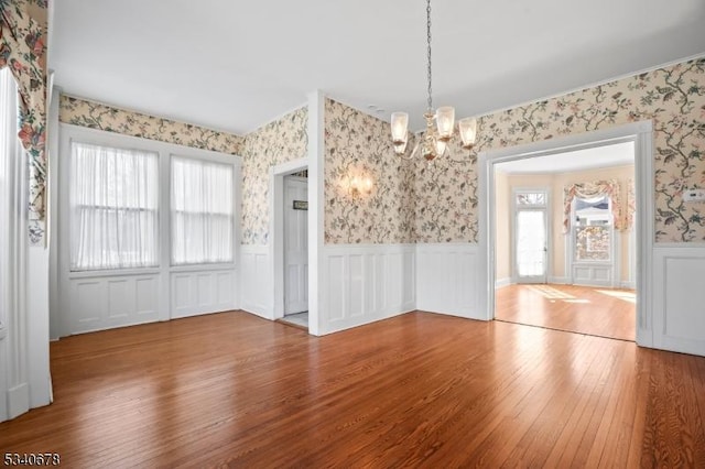 unfurnished dining area featuring wallpapered walls, a notable chandelier, wood finished floors, and wainscoting