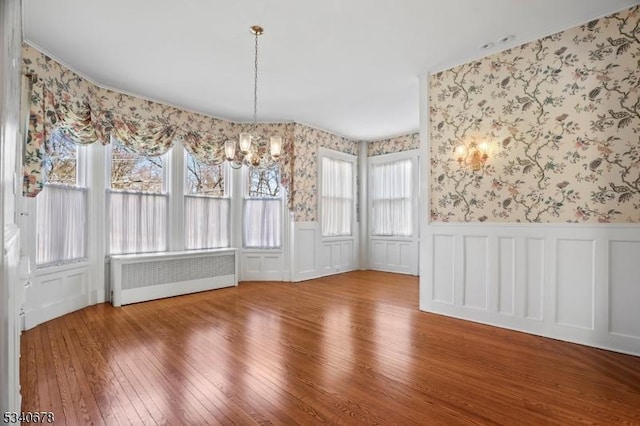 unfurnished dining area featuring hardwood / wood-style flooring, a notable chandelier, wainscoting, radiator heating unit, and wallpapered walls