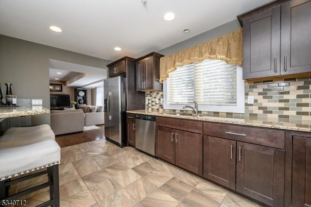 kitchen with a sink, dark brown cabinets, appliances with stainless steel finishes, light stone countertops, and tasteful backsplash