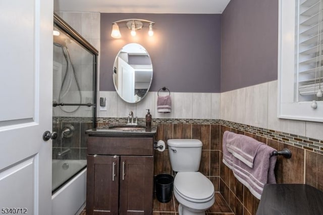 bathroom featuring wainscoting, toilet, combined bath / shower with glass door, vanity, and tile walls