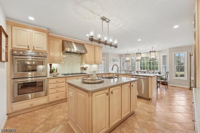 kitchen with stainless steel appliances, light brown cabinetry, wall chimney exhaust hood, and a kitchen island with sink