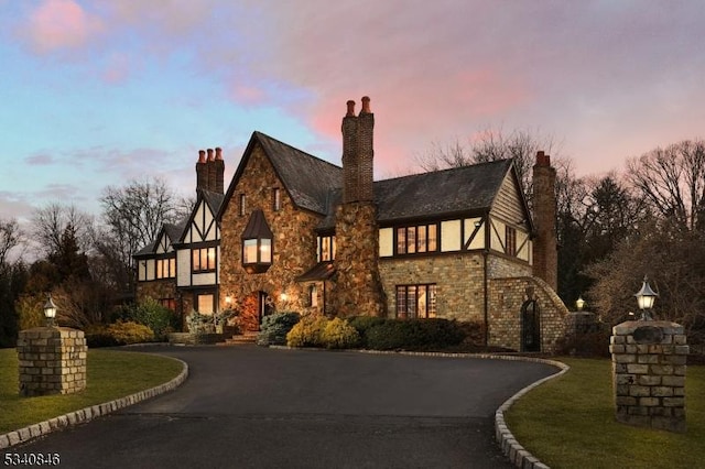 tudor-style house with driveway, a chimney, stone siding, and a lawn