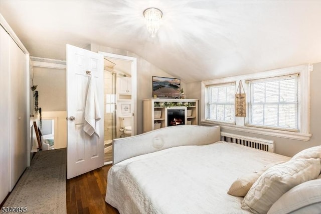 bedroom with radiator heating unit, wood finished floors, ensuite bathroom, a lit fireplace, and vaulted ceiling