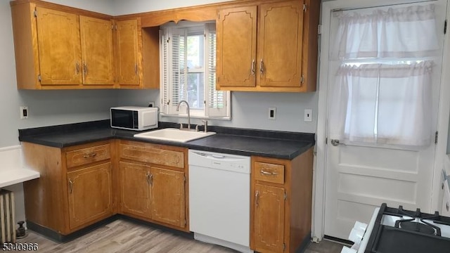 kitchen with dark countertops, a sink, white appliances, brown cabinetry, and light wood-style flooring