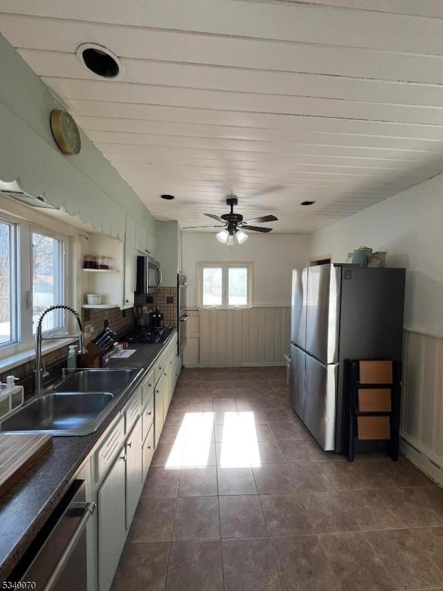 kitchen with a wainscoted wall, stainless steel appliances, dark countertops, a sink, and dark tile patterned floors