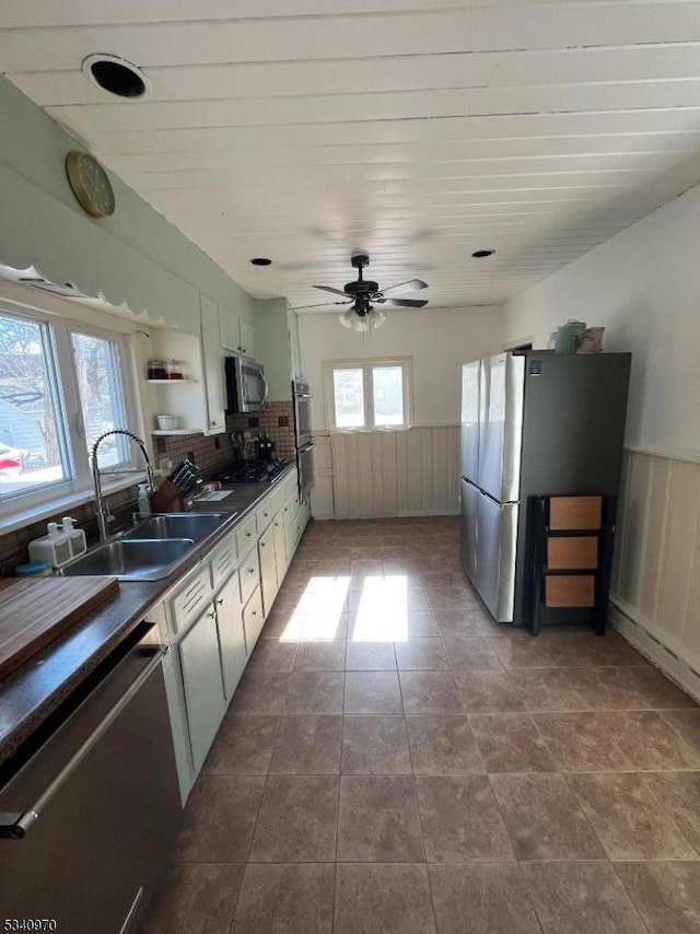 kitchen featuring dark countertops, a wainscoted wall, appliances with stainless steel finishes, open shelves, and a sink