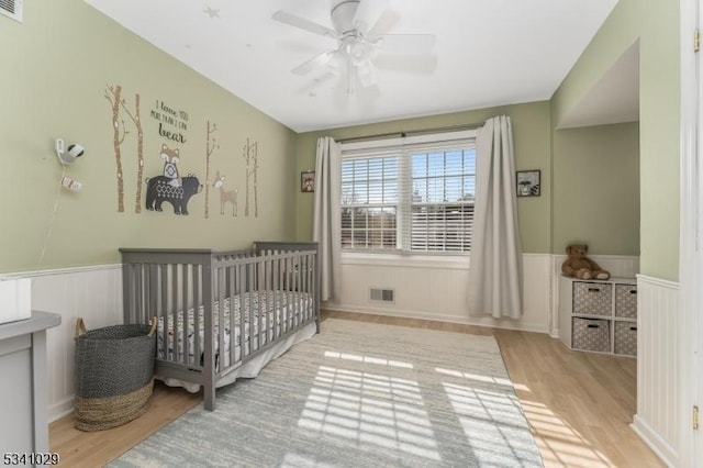 bedroom with a wainscoted wall, a nursery area, visible vents, and wood finished floors