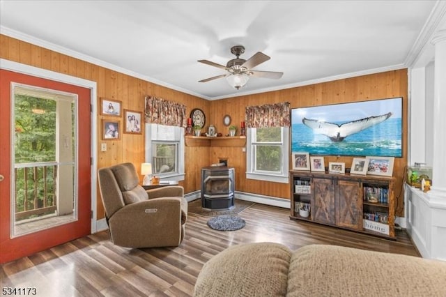 living area with a baseboard radiator, wood finished floors, a ceiling fan, a wood stove, and crown molding