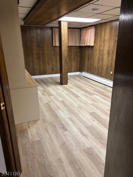 finished below grade area with a baseboard heating unit, light wood-style floors, wood walls, and a drop ceiling