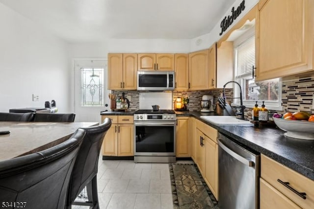 kitchen featuring stainless steel appliances, dark countertops, a wealth of natural light, light brown cabinetry, and a sink