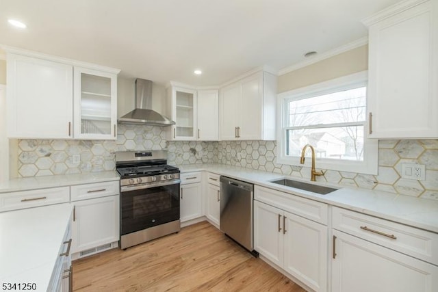 kitchen featuring wall chimney range hood, stainless steel appliances, light wood-style floors, white cabinetry, and a sink