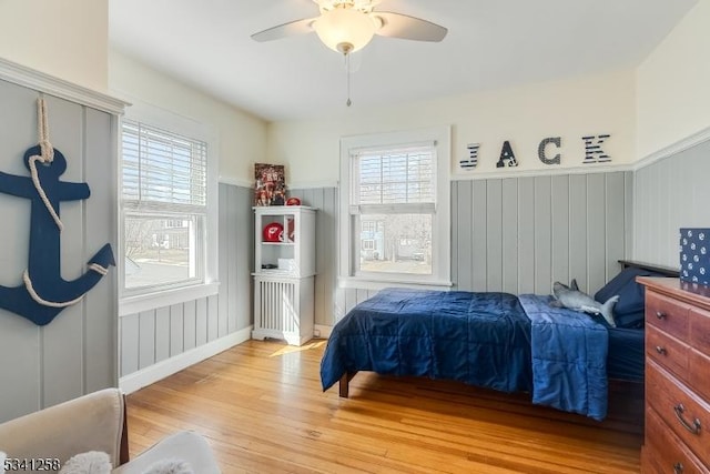 bedroom with a wainscoted wall, multiple windows, a ceiling fan, and wood finished floors