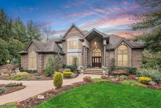 french country home featuring a shingled roof, brick siding, and a yard